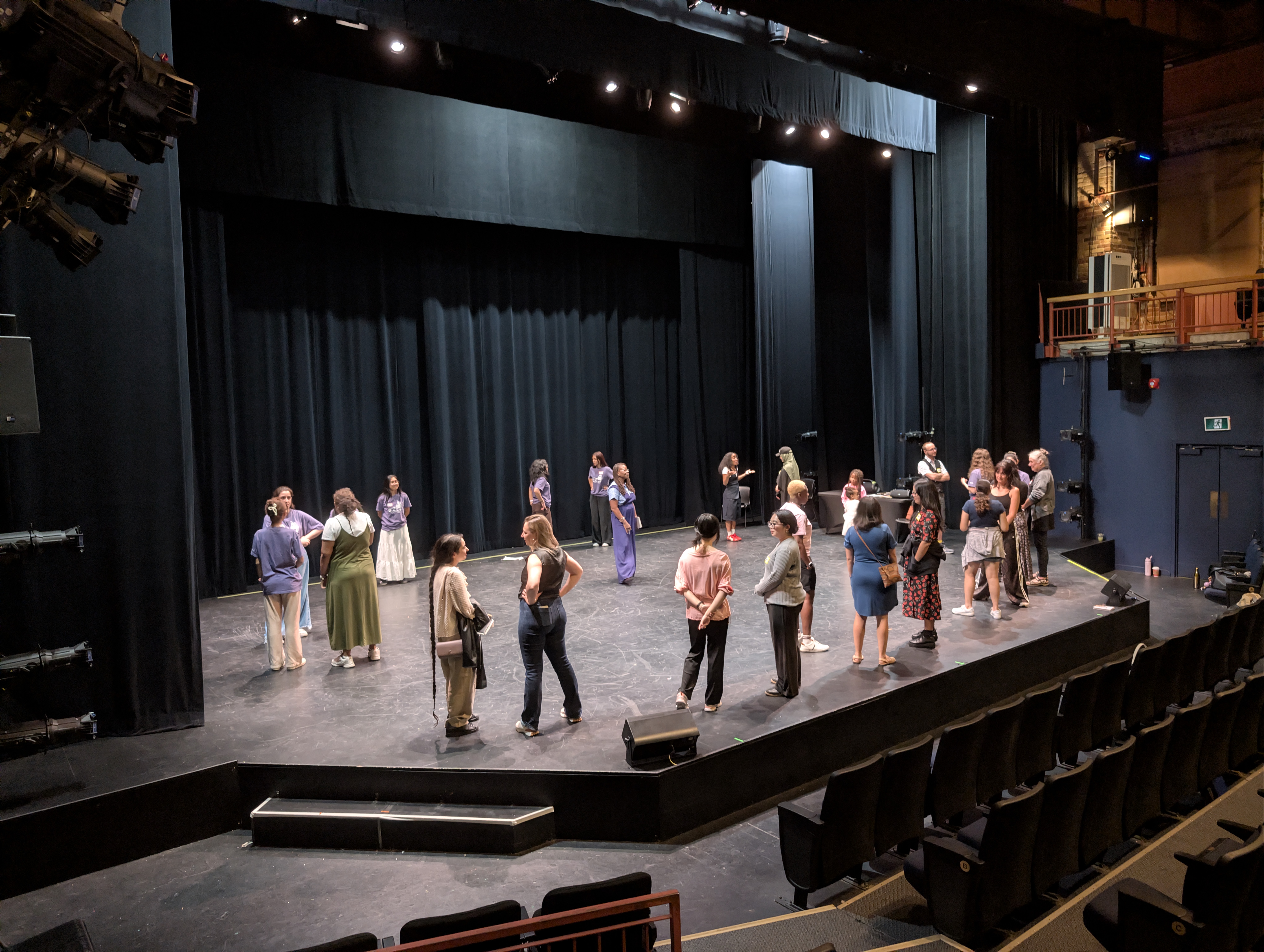 A diverse group of performers gathers on stage in a theater, engaged in conversation and preparation for a rehearsal. Black curtains backdrop the scene.