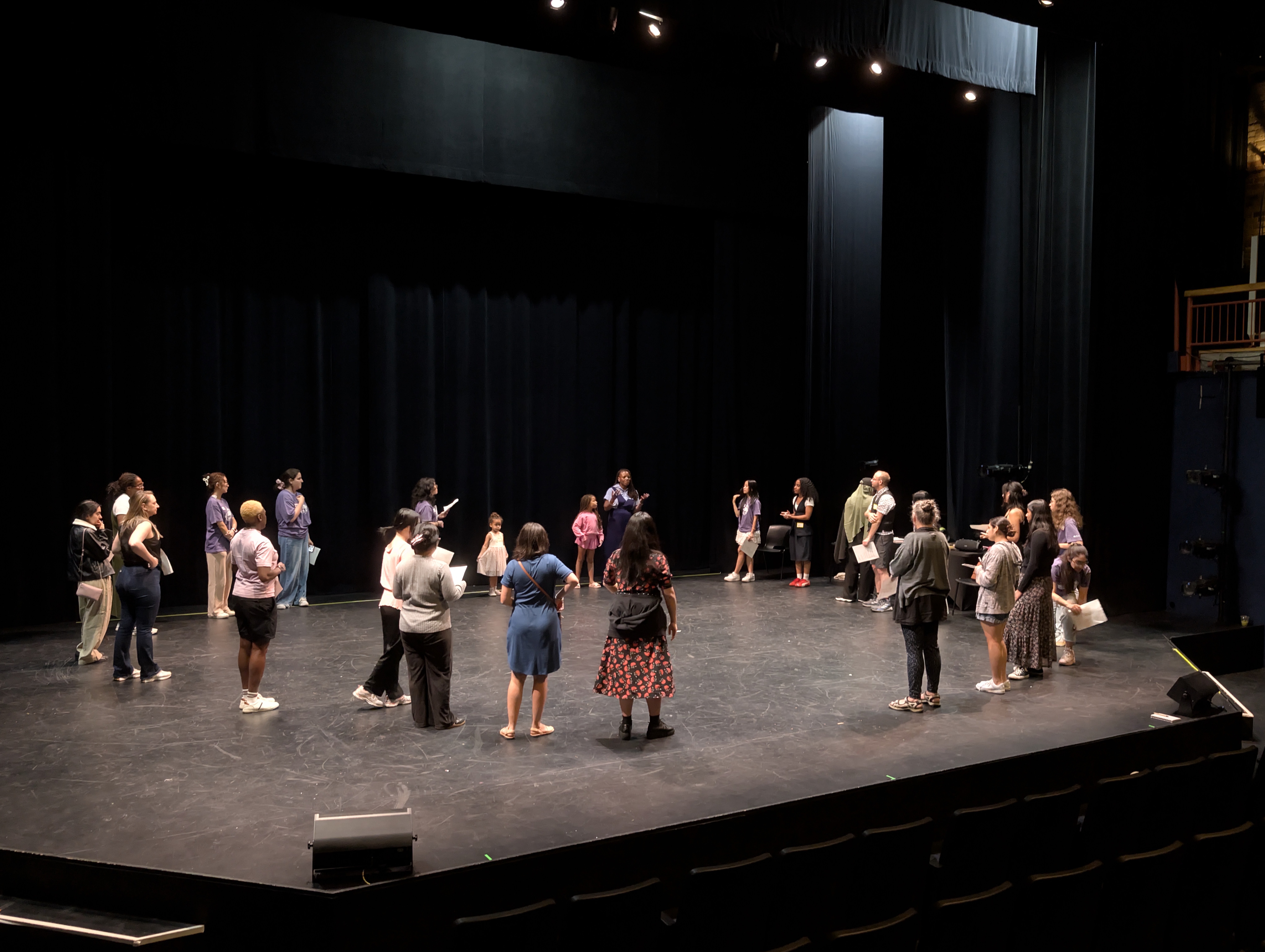 A diverse group of performers rehearses on stage, preparing for a production. The setting features a black curtain backdrop and stage lights.