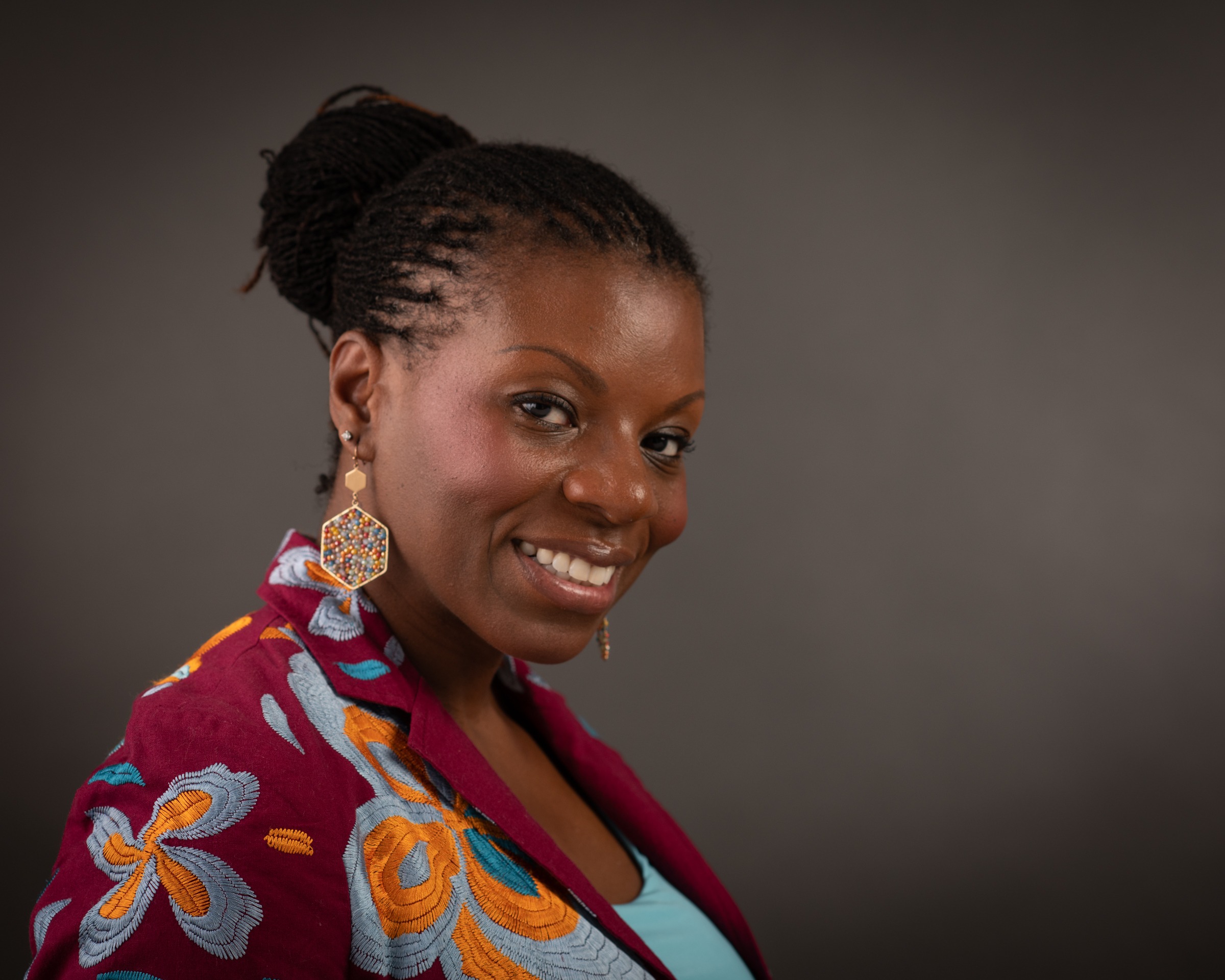 Smiling woman with braided hair, wearing colorful earrings and a floral jacket, poses against a dark background. Radiates confidence and style.