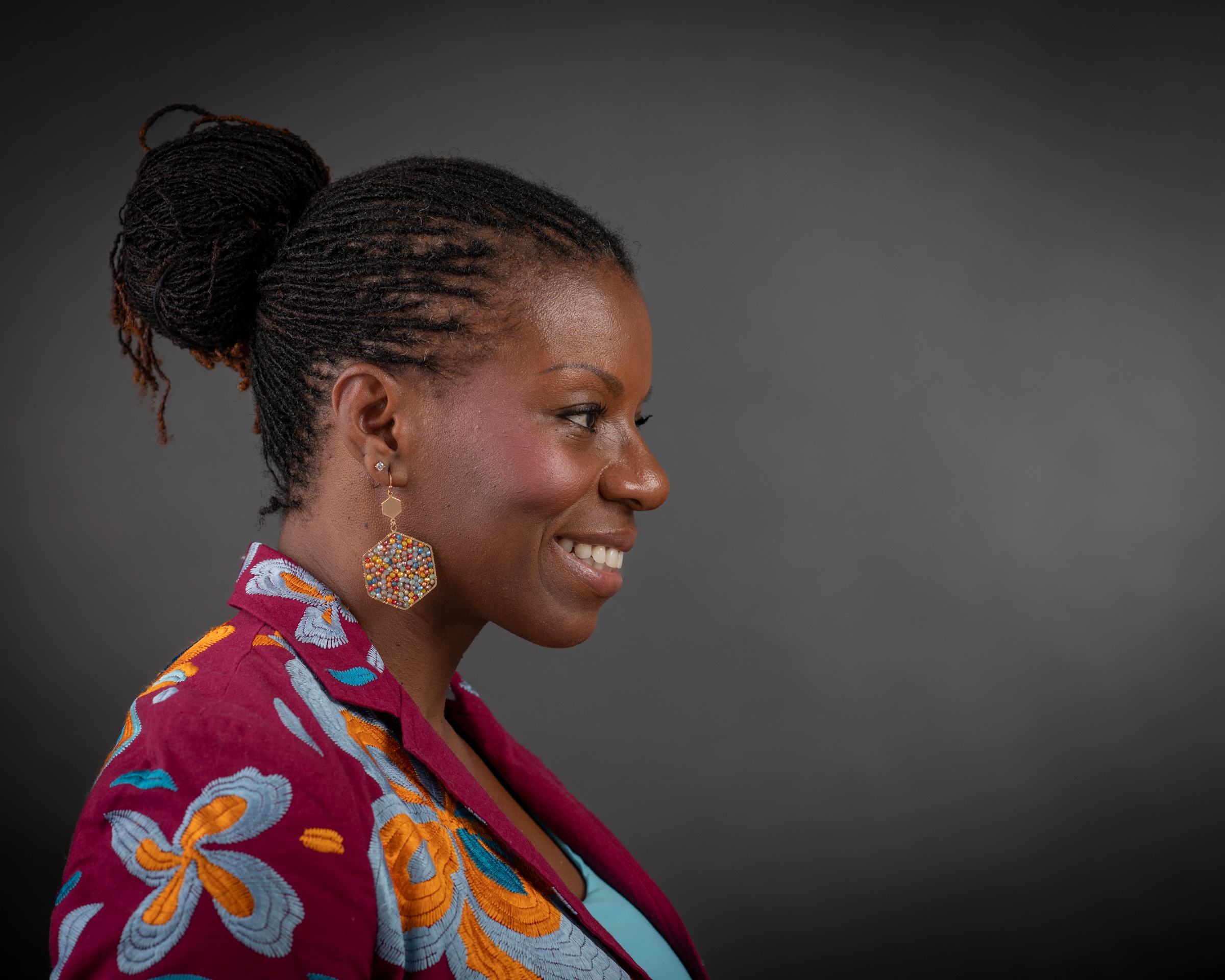 A smiling woman with braided hair worn in a bun, adorned with colorful earrings, wears an embroidered maroon jacket. The background is dark.