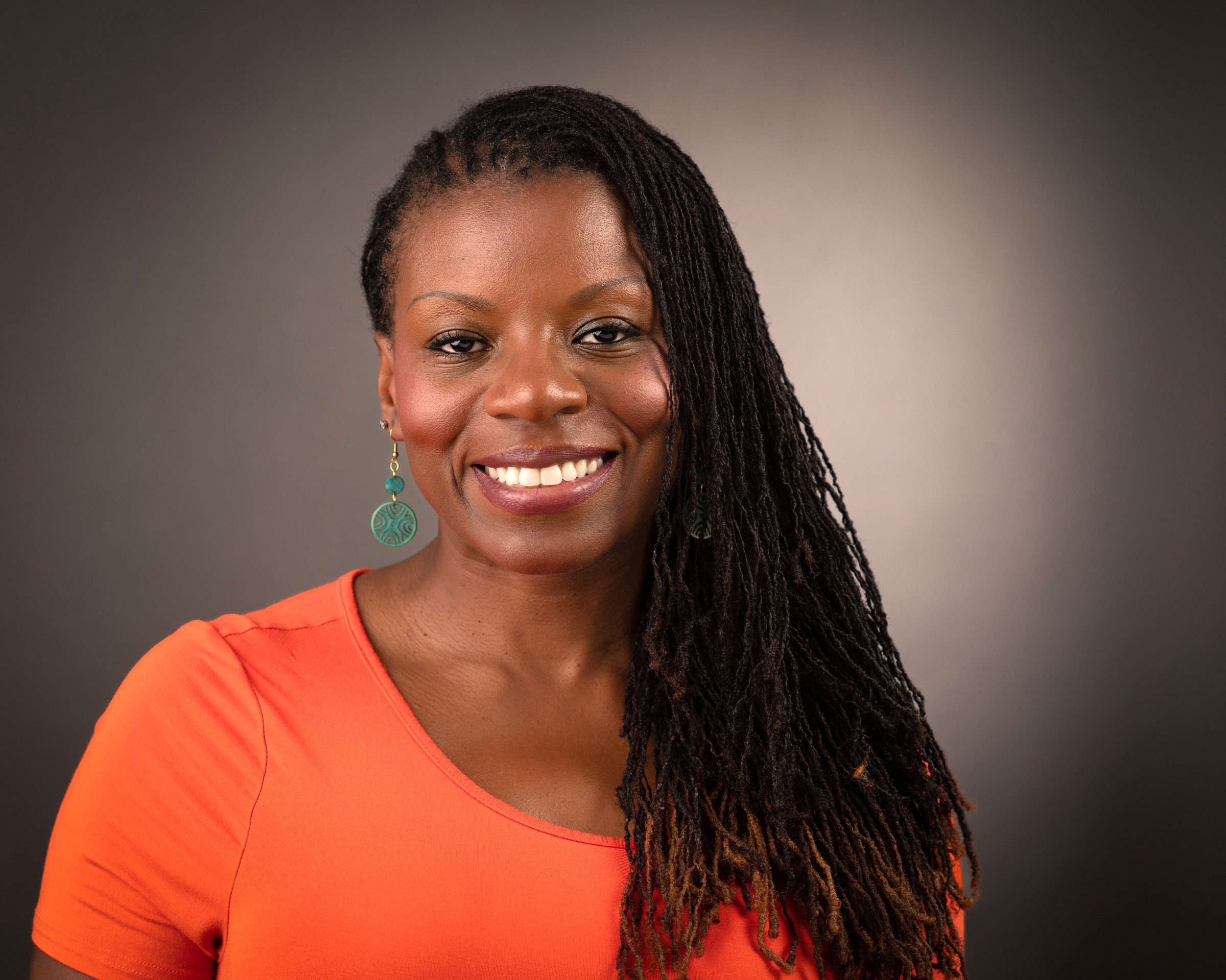 Smiling woman with long, curly hair and teal earrings wears an orange top against a neutral background, conveying warmth and approachability.