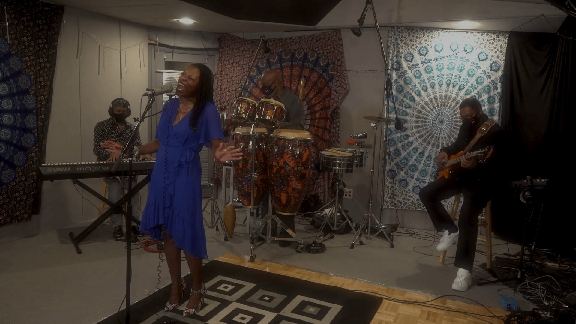 A singer in a blue dress performs passionately on stage, accompanied by musicians playing keyboard, congas, and guitar. Colorful tapestries decorate the backdrop, creating an engaging atmosphere.