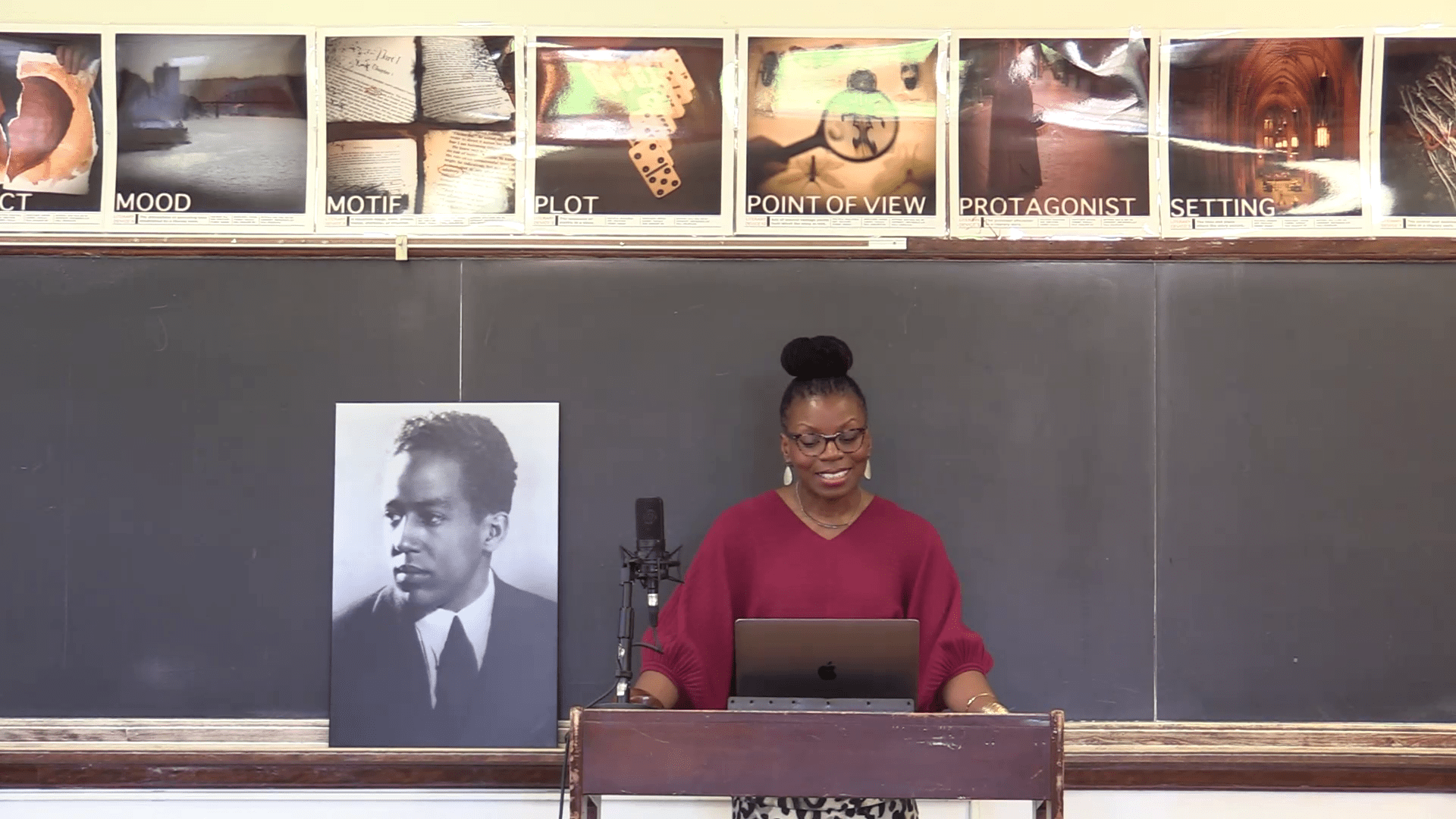 A woman stands at a podium in a classroom, with a laptop and a microphone, smiling. A portrait of a historical figure is displayed nearby, contributing to the lesson on literary elements.