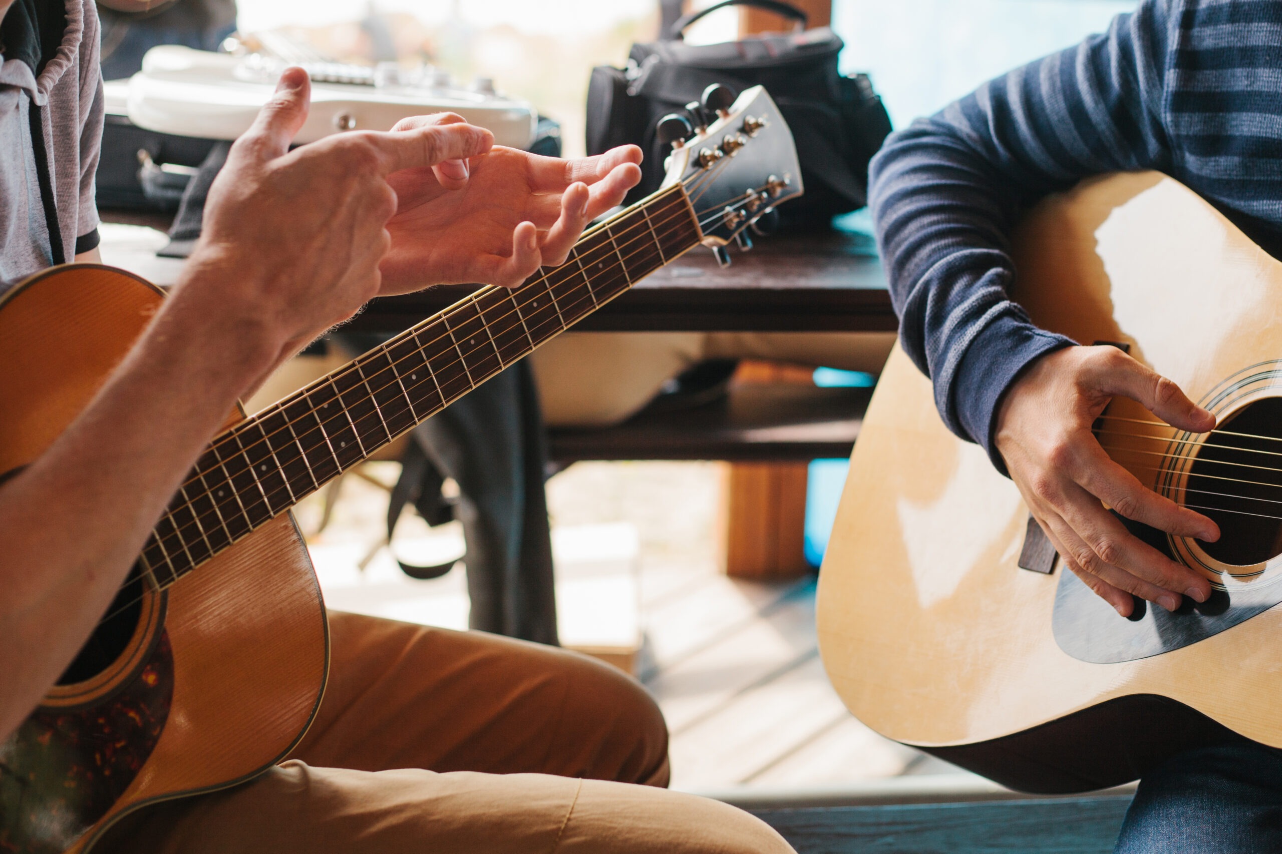 Two musicians play acoustic guitars, with one gesturing to illustrate a point. Natural light filters through a nearby window, creating a relaxed atmosphere.